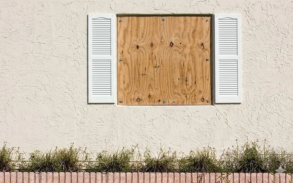 Plywood hurricane panel fully installed and anchored over window on stucco wall with screws visible around perimeter